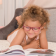 An image of a child laying on a bed and reading a book with an Amazon Kids smart speaker in the background